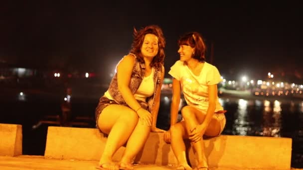 Two pretty ladies sitting at night pier, ocean and boats on background, yellow lights around. Girls talking and smiling, feeling happy at their journey on Bali island, Indonesia. 1080p, 50 fps.  - Footage, Video