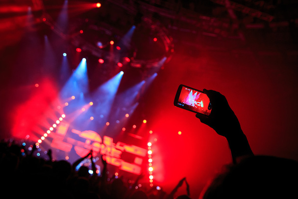 Рука со смартфоном записывает живой музыкальный фестиваль, фотографирует концертную сцену - Фото, изображение
