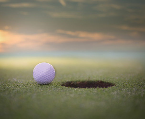 Мяч для гольфа сидит на поле для гольфа, зеленый рядом с лункой. с красивым закатом
. - Фото, изображение