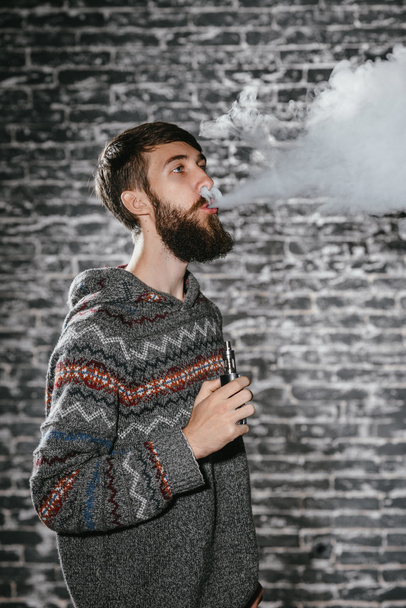 Mladý muž s plnovousem si cucá elektronickou cigaretu. Vaper hipster kouřový odpařovač. - Fotografie, Obrázek