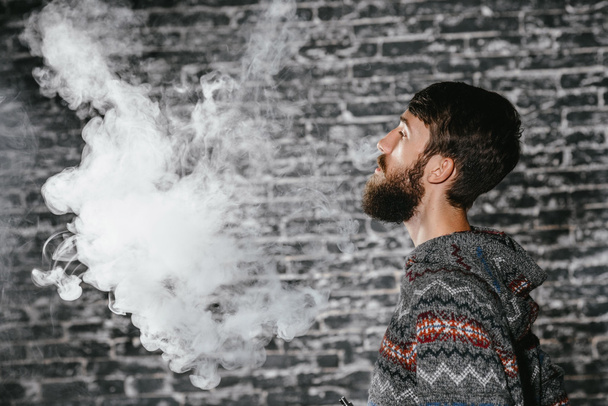 Молодий чоловік з бородатим парінням електронної сигарети. Найкращий гіпсовий димовий випарник
. - Фото, зображення