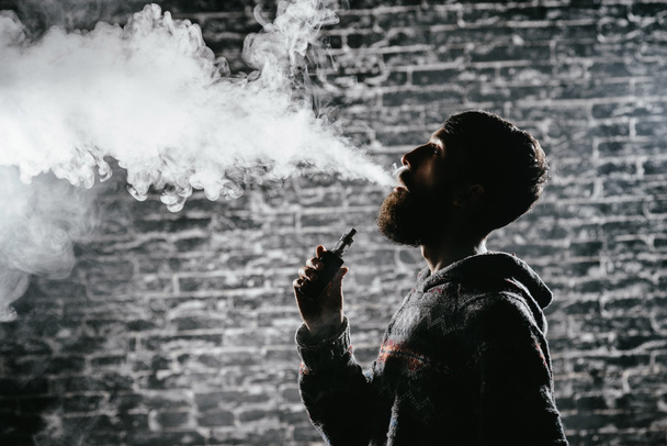 Un joven con barba vapeando un cigarrillo electrónico. Vaporizador de humo Vaper hipster
. - Foto, Imagen