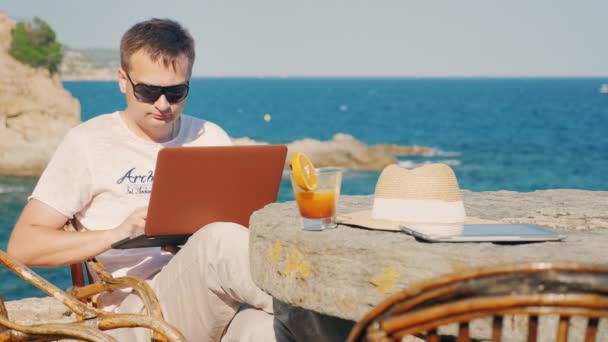 Man in sunglasses work with a laptop. Against the background of the sea at a table in a cafe - Footage, Video