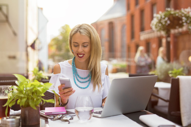 Mujer sentada en la cafetería con portátil y charlando en su teléfono móvil
 - Foto, Imagen