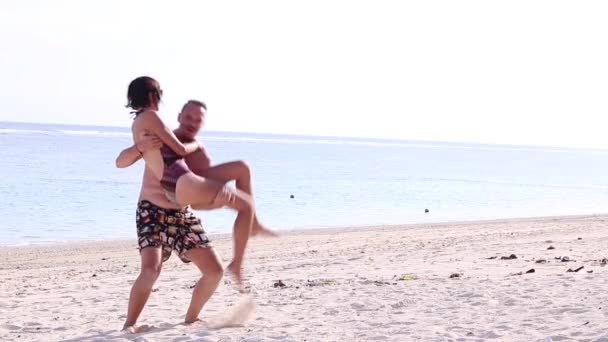 Young man holds and turns his sexy girlfriend on the beach of Bali island, Indonesia. Scene of couple playing on the tropical beach. Happy young guys swirling. Slow motion. - Filmati, video