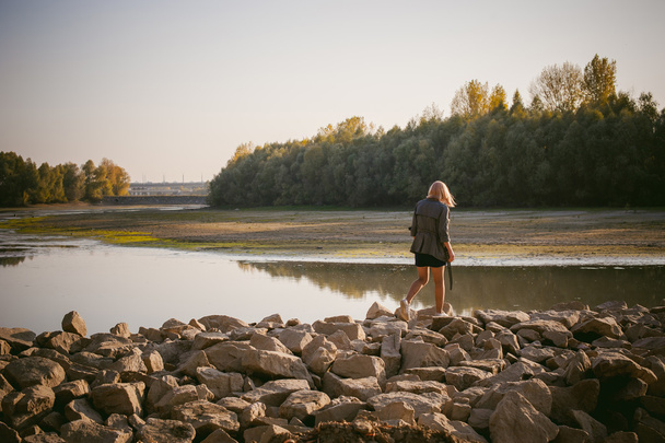 pretty blonde girl in sneakers, nylon stockings and coat. walking on the stone shore of the lake in the last sunshine of the departing summer. - Фото, зображення