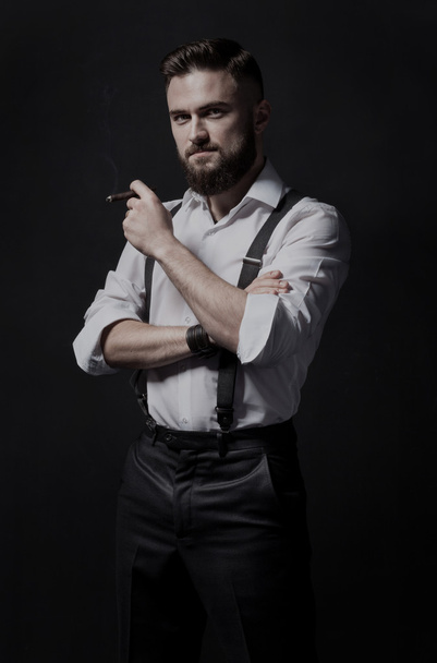 Hombre barbudo joven, elegante y atractivo fumando un cigarro en una habitación oscura. Lleva puesta una camisa blanca y tirantes.
. - Foto, Imagen
