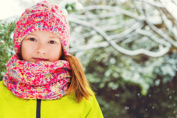 Zimní portrét roztomilá holčička za sněžení, nosí zelený kabát, barevná pletená čepice a šála - Fotografie, Obrázek