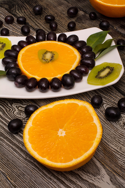 slice of orange, black grapes, kiwi on a white plate, wooden background - Foto, Imagen