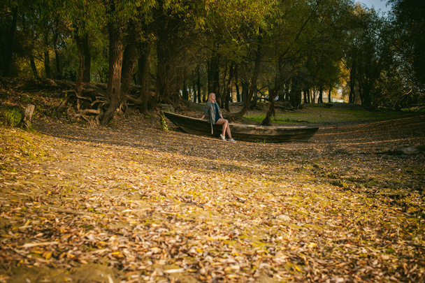 Pretty blonde sitting in a wooden old boat which is on the ground laid by yellow fallen autumn leaves - Foto, immagini