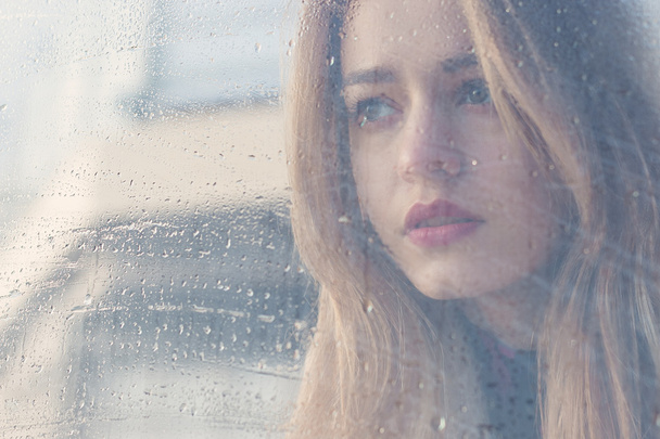 Beautiful sad girl with big eyes in a coat is behind wet glass - Foto, Bild