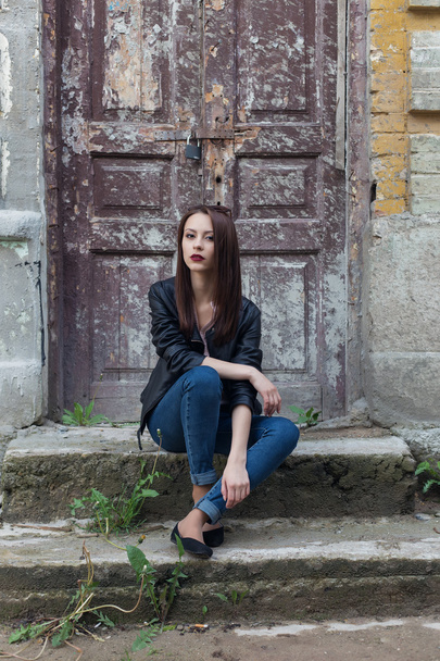красивая милая девушка моды с темными волосами в темных очках в кожаной черной куртке сидит на лестнице к крыльцу старых зданий в городе
 - Фото, изображение