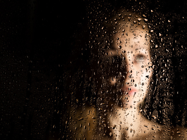 sexy young woman, posing behind transparent glass covered by water drops. melancholy and sad female portrait - Fotó, kép