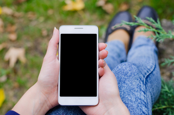 Λευκό κινητό τηλέφωνο στο χέρι μια γυναίκα των επιχειρήσεων νέων hipster στο φόντο πράσινο φυσικό θάμνο και το φθινόπωρο φύλλα μια πέτρινη πλάκα πλάκες επίστρωσης - Φωτογραφία, εικόνα