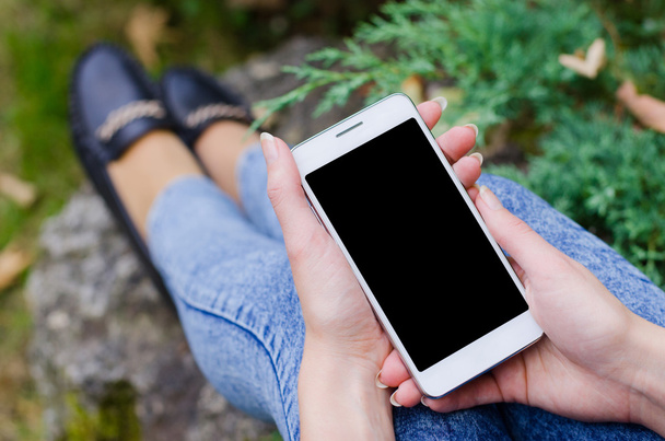 Telefone celular branco na mão uma jovem mulher de negócios hipster no fundo do arbusto natural verde e folhas de outono de uma laje de pedra lajes de pavimentação
 - Foto, Imagem
