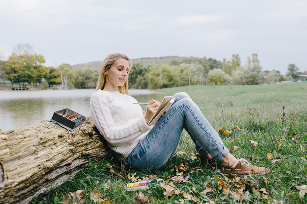 Pretty smiling young woman in white sweater and blue jeans draws in a notebook with colored pencils sitting on the grass and leaned into the trunk of a dry tree on a background of lake - Photo, Image