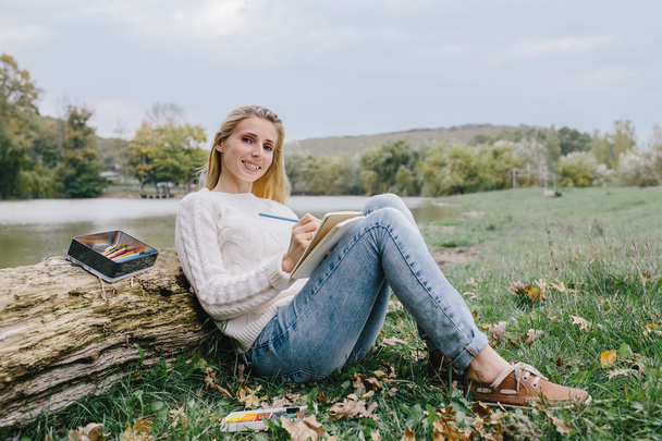 Bella giovane donna sorridente in maglione bianco e jeans blu disegna in un quaderno con matite colorate seduto sull'erba e appoggiato nel tronco di un albero secco su uno sfondo di lago
 - Foto, immagini