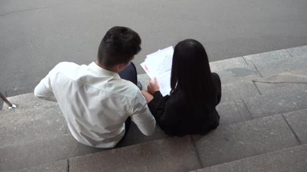 Young Asian Business Woman Female and Caucasian Male Use Look at Documents Sit on Office Building Stairs - Video