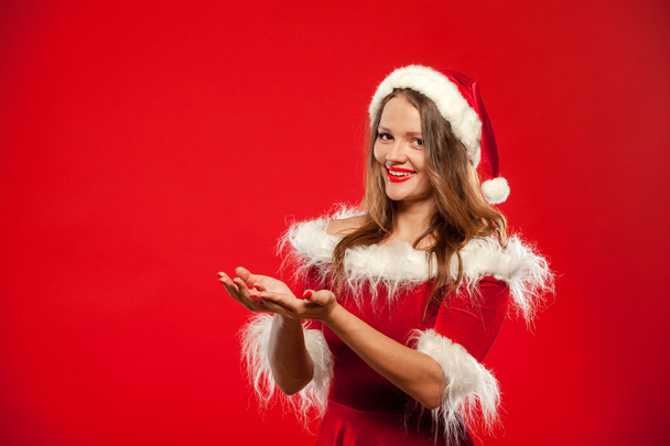 christmas, x-mas, winter, happiness concept - smiling woman in santa helper hat with gift box, holding hands in front of him, offering a , over red background. - Zdjęcie, obraz
