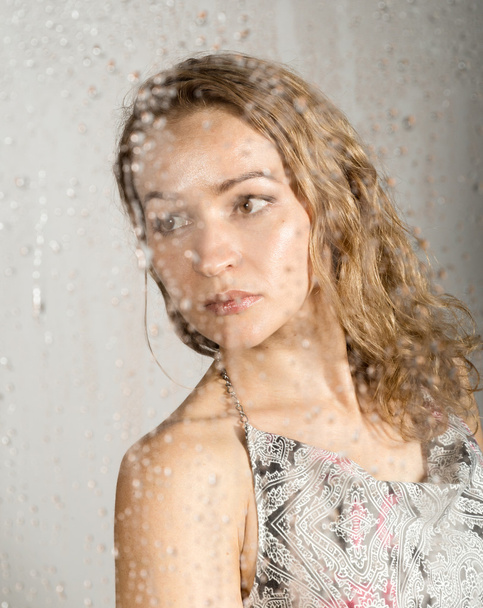 sexy young woman, posing behind transparent glass covered by water drops. melancholy and sad female portrait - 写真・画像