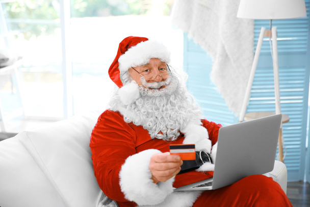 Santa Claus con tarjeta de crédito y portátil en el sofá en casa
 - Foto, Imagen