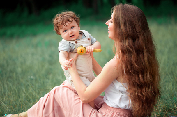 Genç güzel annem yeşil çim karşı küçük oğluyla oturuyor. Güneşli bir yaz onun erkek bebek ile mutlu bir kadın. Çayır üzerinde yürüme aile. - Fotoğraf, Görsel