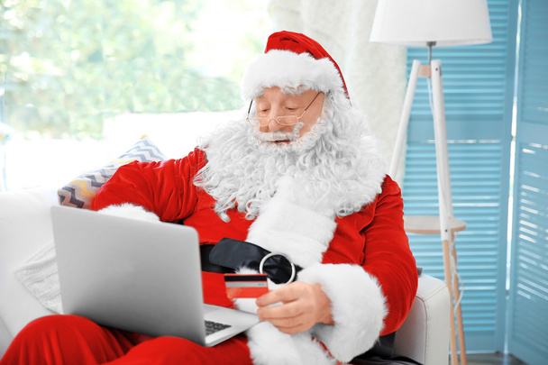 Santa Claus with credit card and laptop on couch at home - Foto, imagen