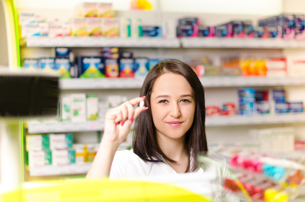 Gyógyszerész gyógyszertár nő mutat egy pirulát. Gyógyszertár-gyógyszertár a háttérben. Csinos, fiatal laborant.  - Fotó, kép