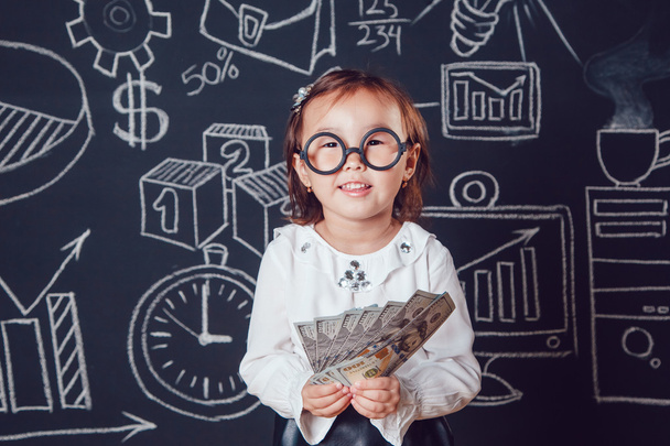 Маленькая умная девочка в очках держит деньги на фоне стены с деловой или школьной фотографией
 - Фото, изображение