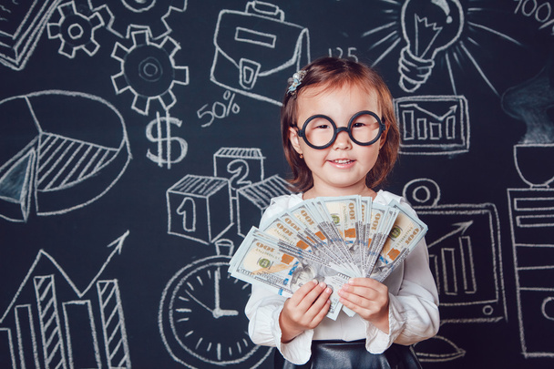 Маленькая умная девочка в очках держит деньги на фоне стены с деловой или школьной фотографией
 - Фото, изображение