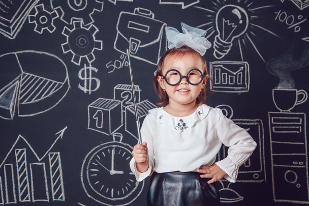 Маленькая умная девочка в очках держит указатель на темном фоне с деловой или школьной картинкой
 - Фото, изображение
