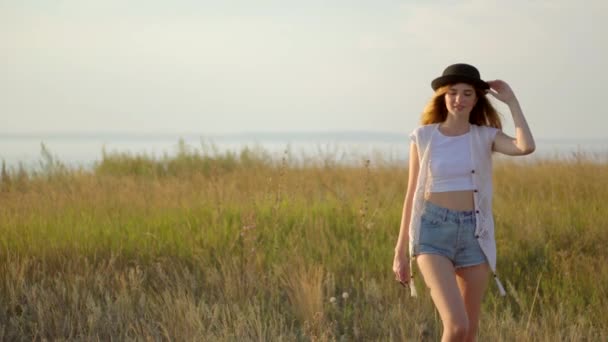 Молода красива дівчина модель в капелюсі ходить і позує на лузі біля річки
 - Кадри, відео