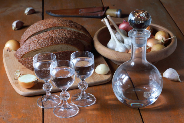Граф и три стакана водки, стоящие на деревянном столе рядом с ломтиками хлеба и тарелкой с овощами
. - Фото, изображение