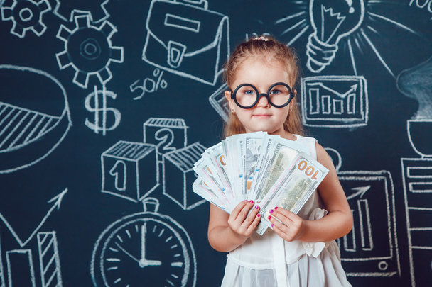The little smart girl in glasses holding money on a background of wall with business or school picture - Photo, Image