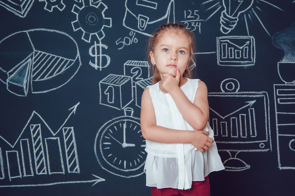Το μικρό έξυπνο κορίτσι που κρατά το πηγούνι της σε φόντο τοίχου με επαγγελματική ή σχολική εικόνα - Φωτογραφία, εικόνα