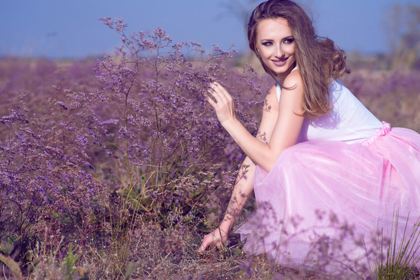 Joven mujer elegante con el pelo ondulado largo y maquillaje artístico sentado en el campo de flores violetas mirando a un lado y sonriendo
. - Foto, imagen