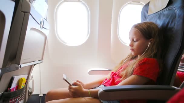 Imádnivaló kislány, aki repülővel utazik az ablak mellett. Gyerek zenét hallgatni és üzenetet küldeni ül közel repülőgép ablak - Felvétel, videó