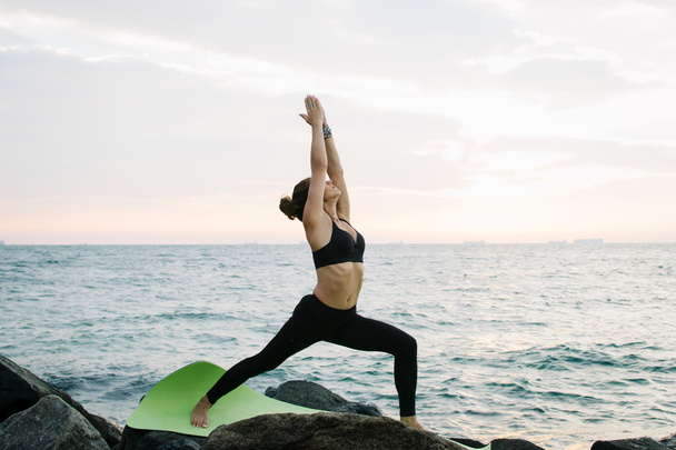 Young woman practicing yoga at rocky seashore at sunrise or sunset - Foto, imagen