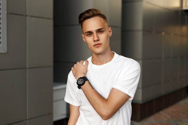 Joven chico guapo en una camiseta blanca posando cerca de la pared gris moderna
. - Foto, imagen