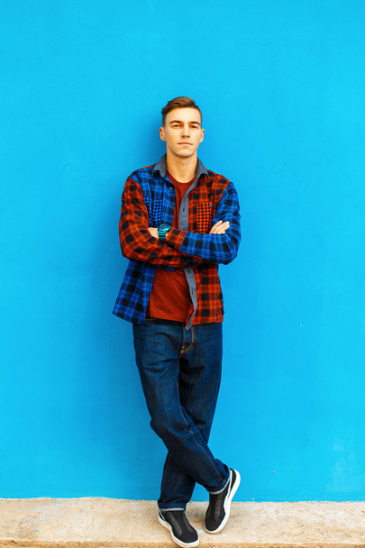 Elegante hombre guapo en una camisa de moda, zapatillas de deporte y jeans se encuentra cerca de una pared azul brillante
. - Foto, imagen