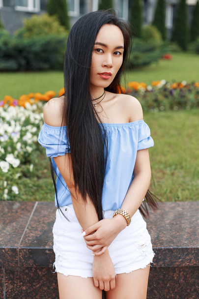 Beautiful young Asian girl in a summer blue blouse and white shorts is posing near the alley of flowers. - Photo, Image