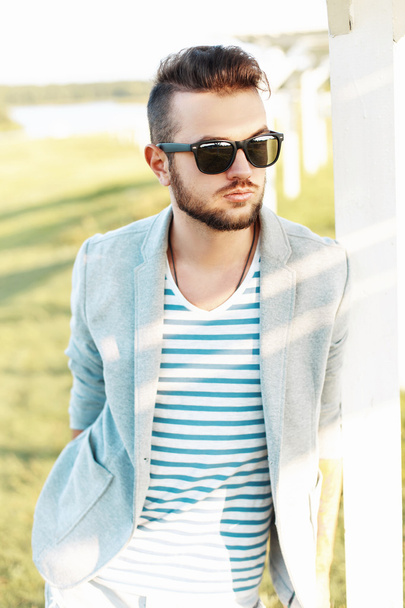 Beau hipster homme vêtu d'une veste, d'un T-shirt d'été et de lunettes de soleil par une journée ensoleillée
. - Photo, image