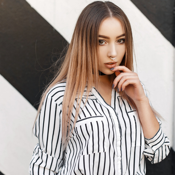 Beautiful modern style girl with earrings in the nose in a fashionable striped blouse near the wall - Photo, Image