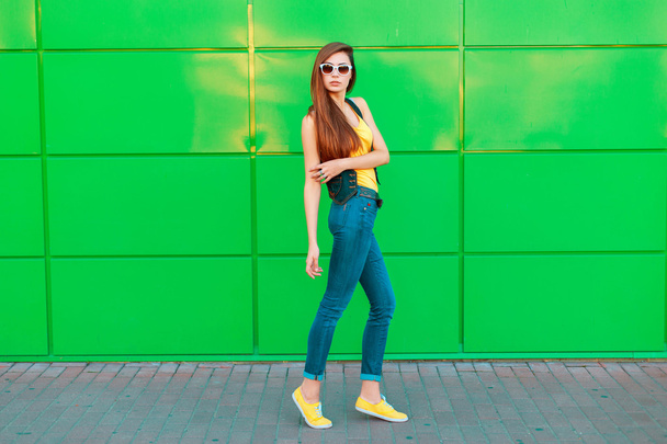 Молода красива дівчина в сонцезахисних окулярах в жовтій футболці, джинсах і кросівках позує біля зеленої стіни
 - Фото, зображення