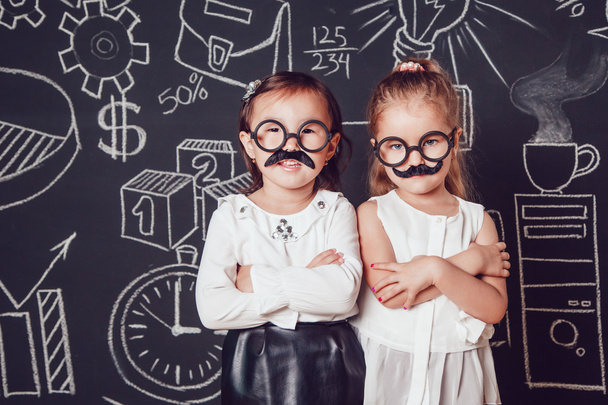 Two little girls as with mustache and glasses standing on dark background of painted wall. Their hands folded - Foto, afbeelding