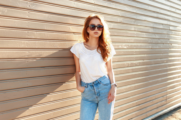 Piękna stylowa dziewczyna w vintage jeans niebieski i biały T-shirt stojący w pobliżu ściany drewniane. - Zdjęcie, obraz