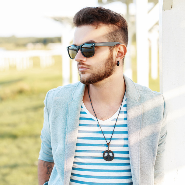 Stylish hipster guy with hairstyle and beard in sunglasses in a trendy jacket resting on the beach in summer sunny day. - Photo, Image