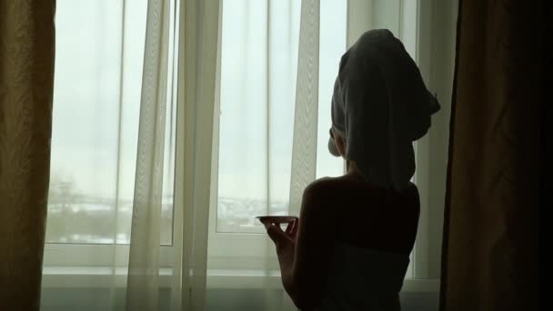 Seductive girl dressed in towel with neat body is holding a cup with hot tea or coffee, standing near the window in her home or hotel room - Metraje, vídeo