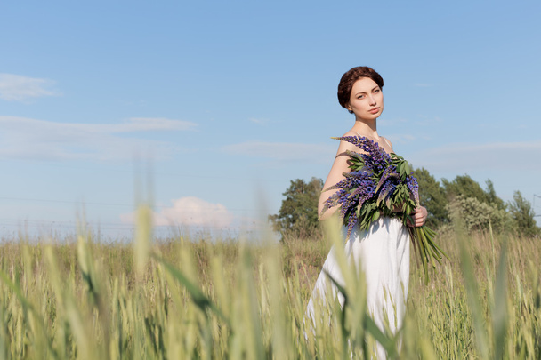 beautiful sweet girl with a plait hair in a white summer sundress walking in a field with a bouquet of purple lupine flowers - Photo, image