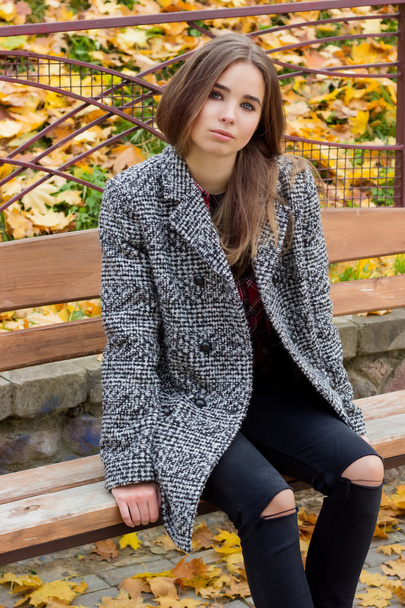 krásná mladá dívka s velkou podzimní smutnýma očima kabát a roztrhl černé džíny sedí na lavičce v podzimním parku - Fotografie, Obrázek